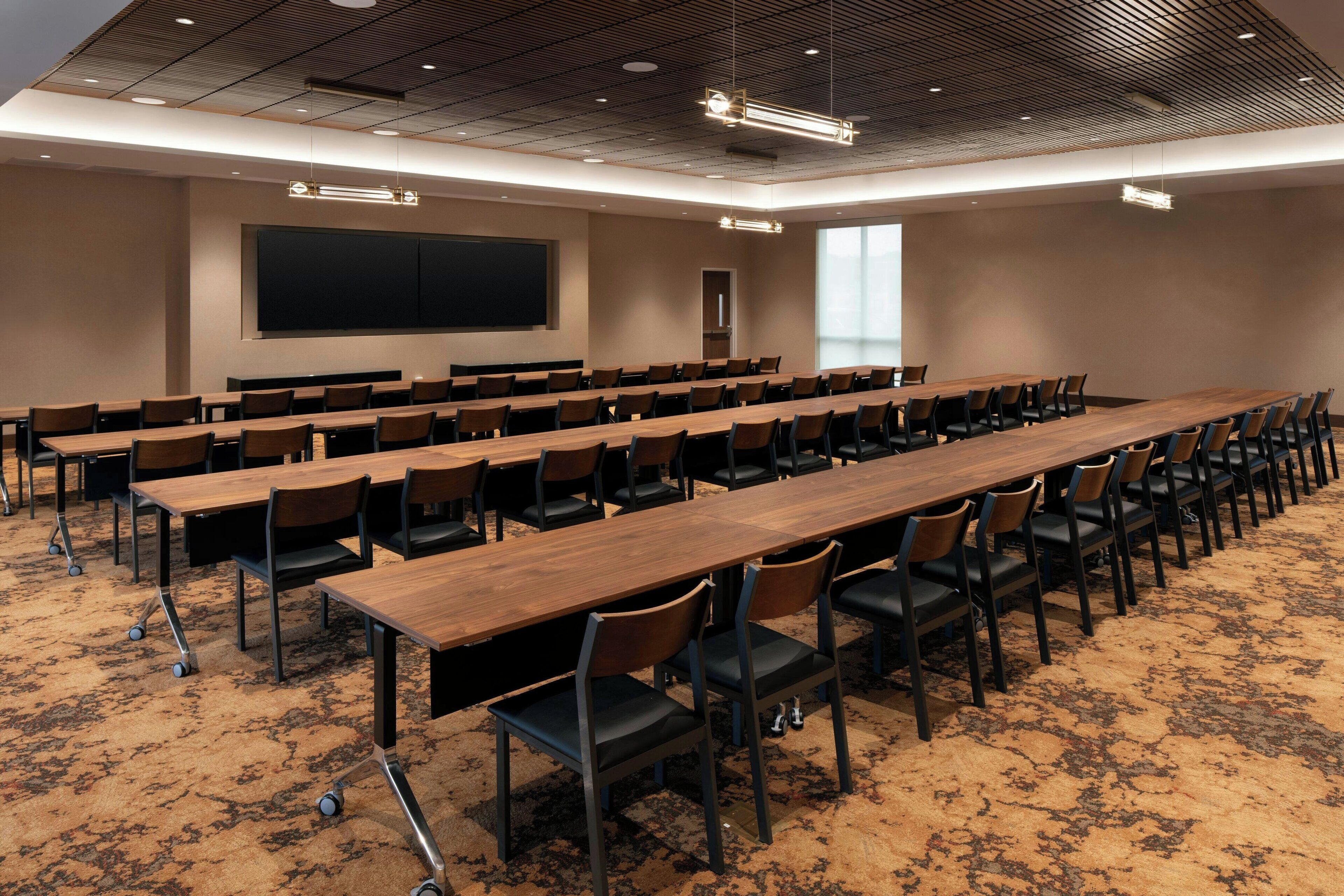 Un salón de conferencias con mesas largas y sillas, una pantalla de proyector y un ventanal grande.