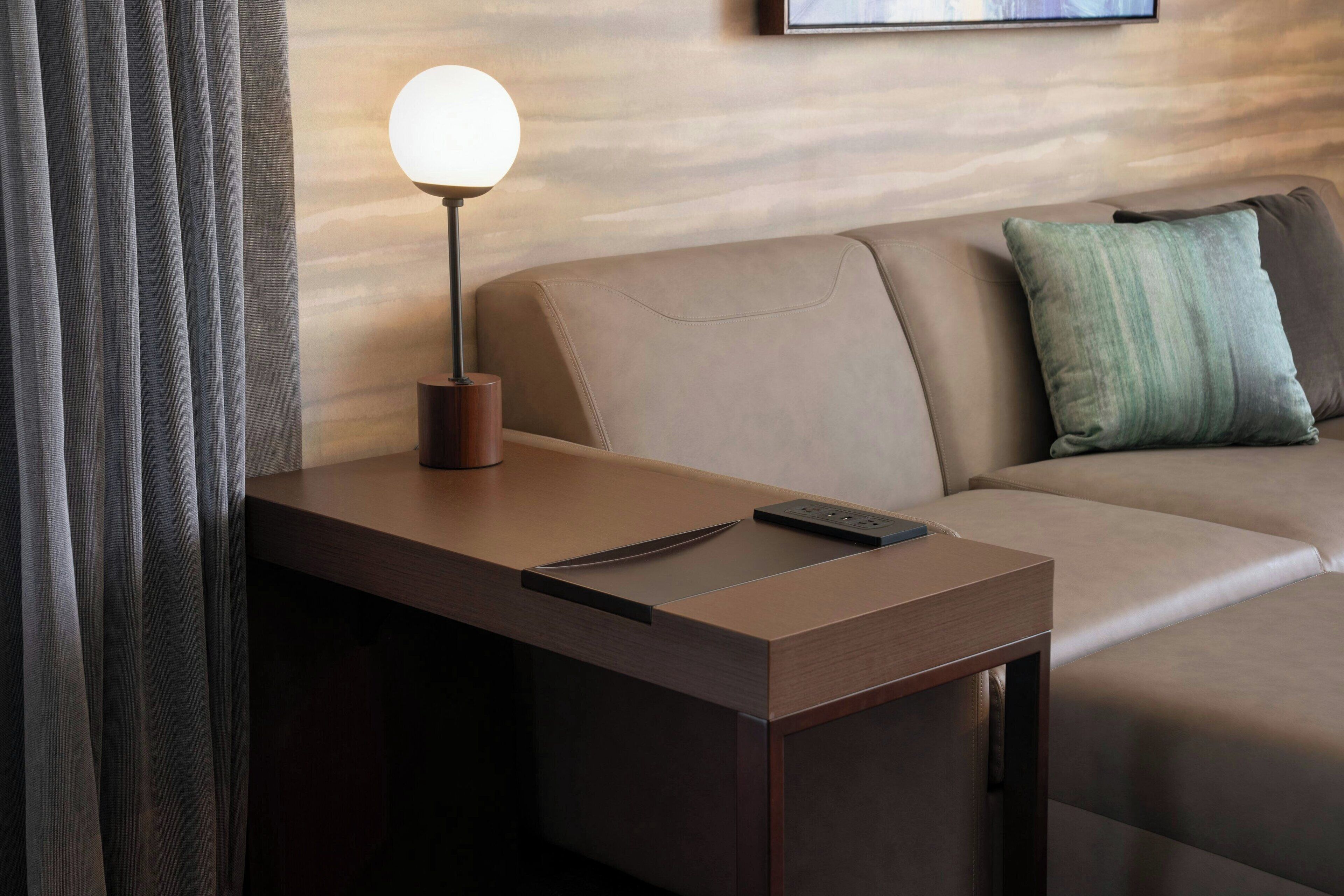 A modern hotel room with a wooden desk, a lamp, a remote control, and a sofa with a green pillow.