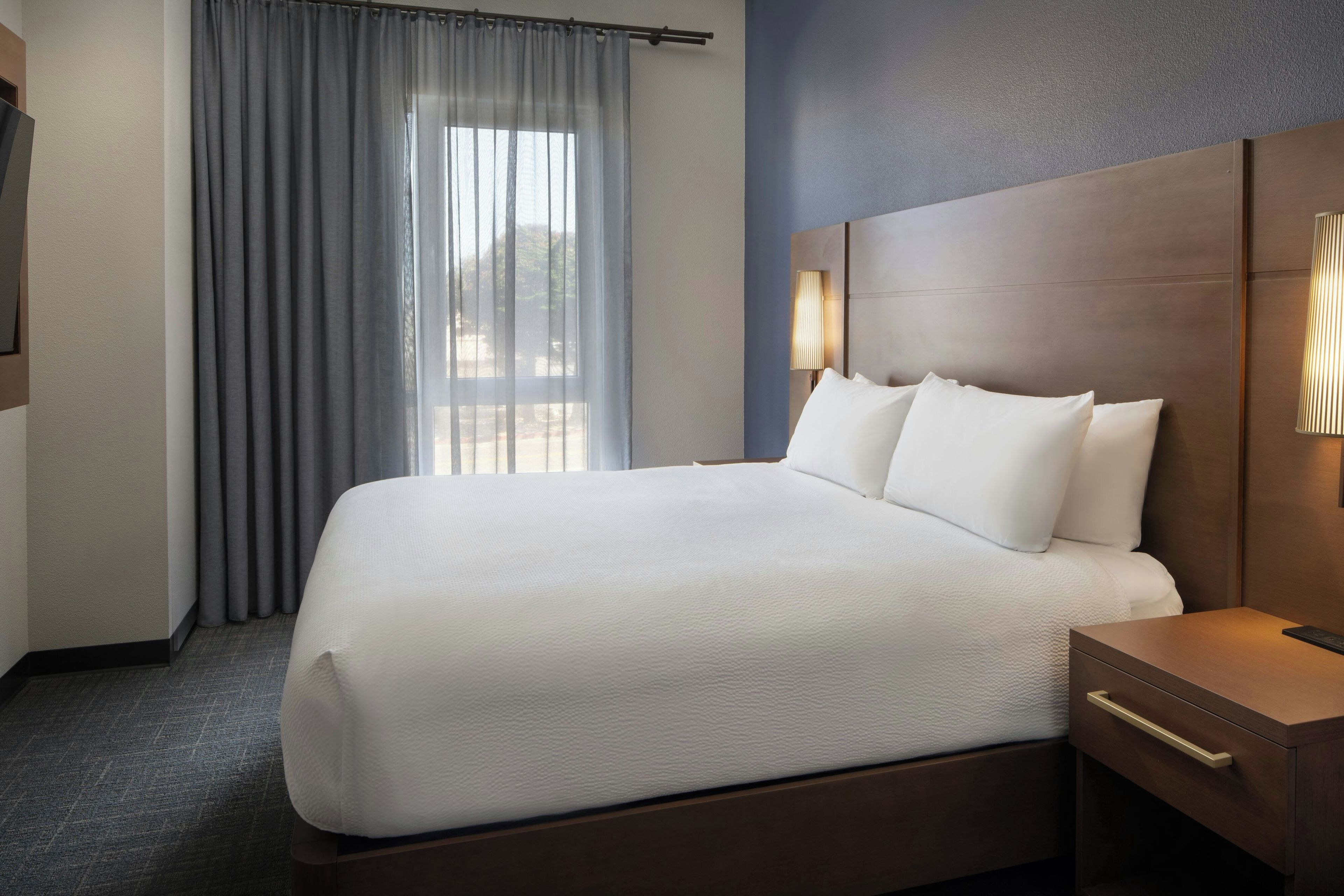 A hotel room with a large bed, a nightstand, a lamp, and a view of the outdoors through the window.