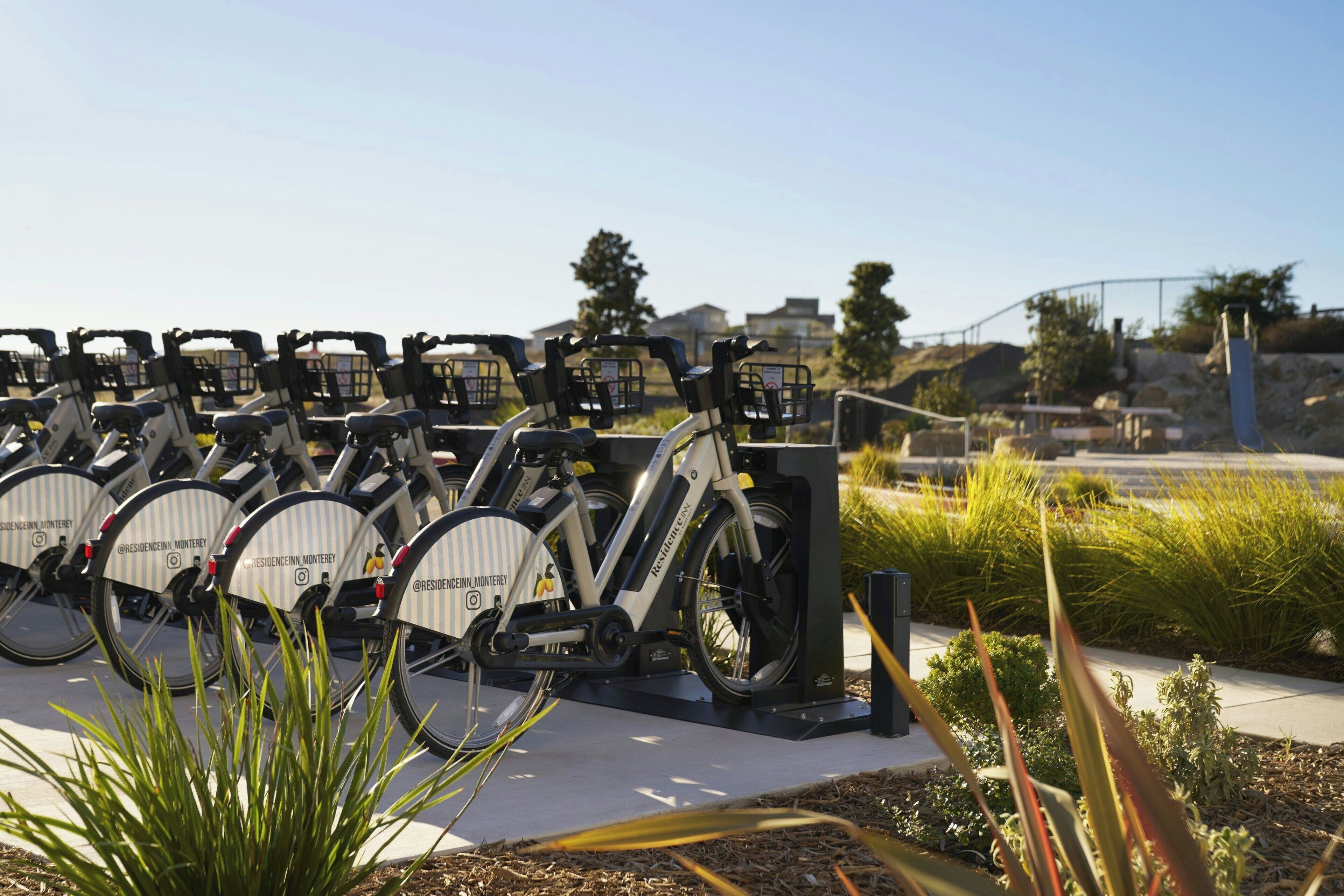 Una hilera de bicicletas estáticas con calcomanías de marca, estacionadas en un área exterior con cielo despejado y algo de vegetación al fondo.