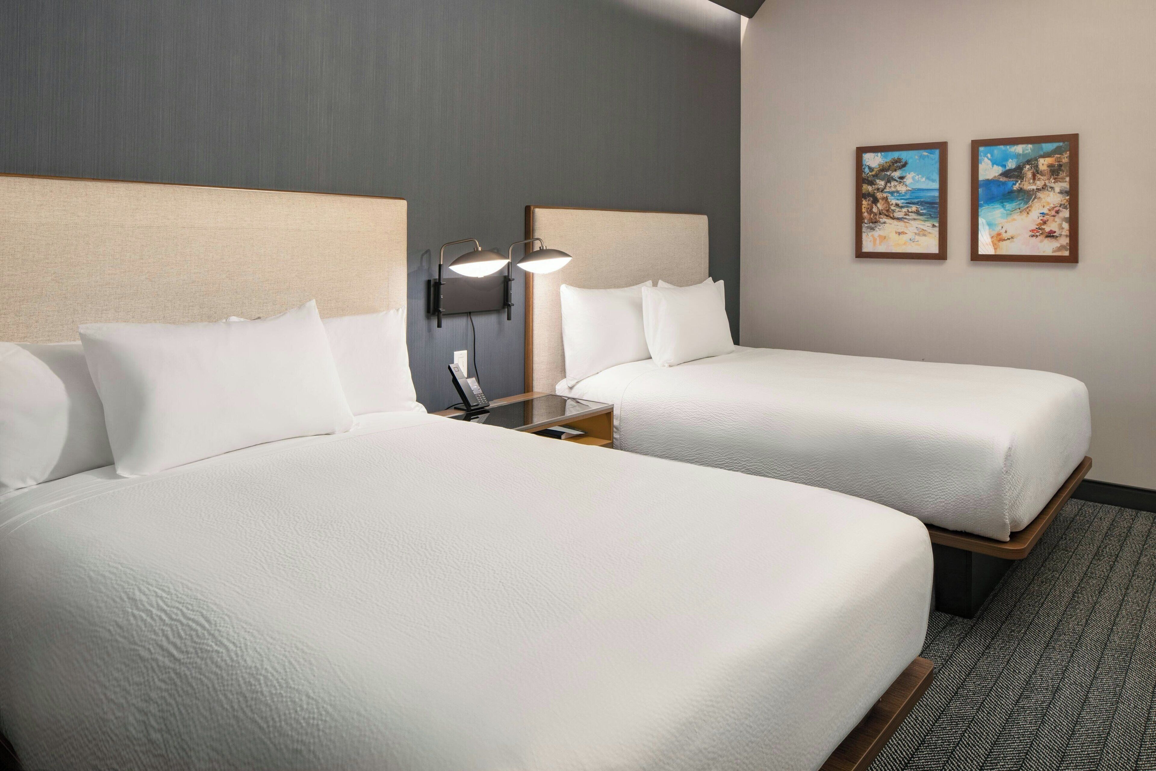 A hotel room with two beds, a nightstand with a lamp, and two framed pictures on the wall.