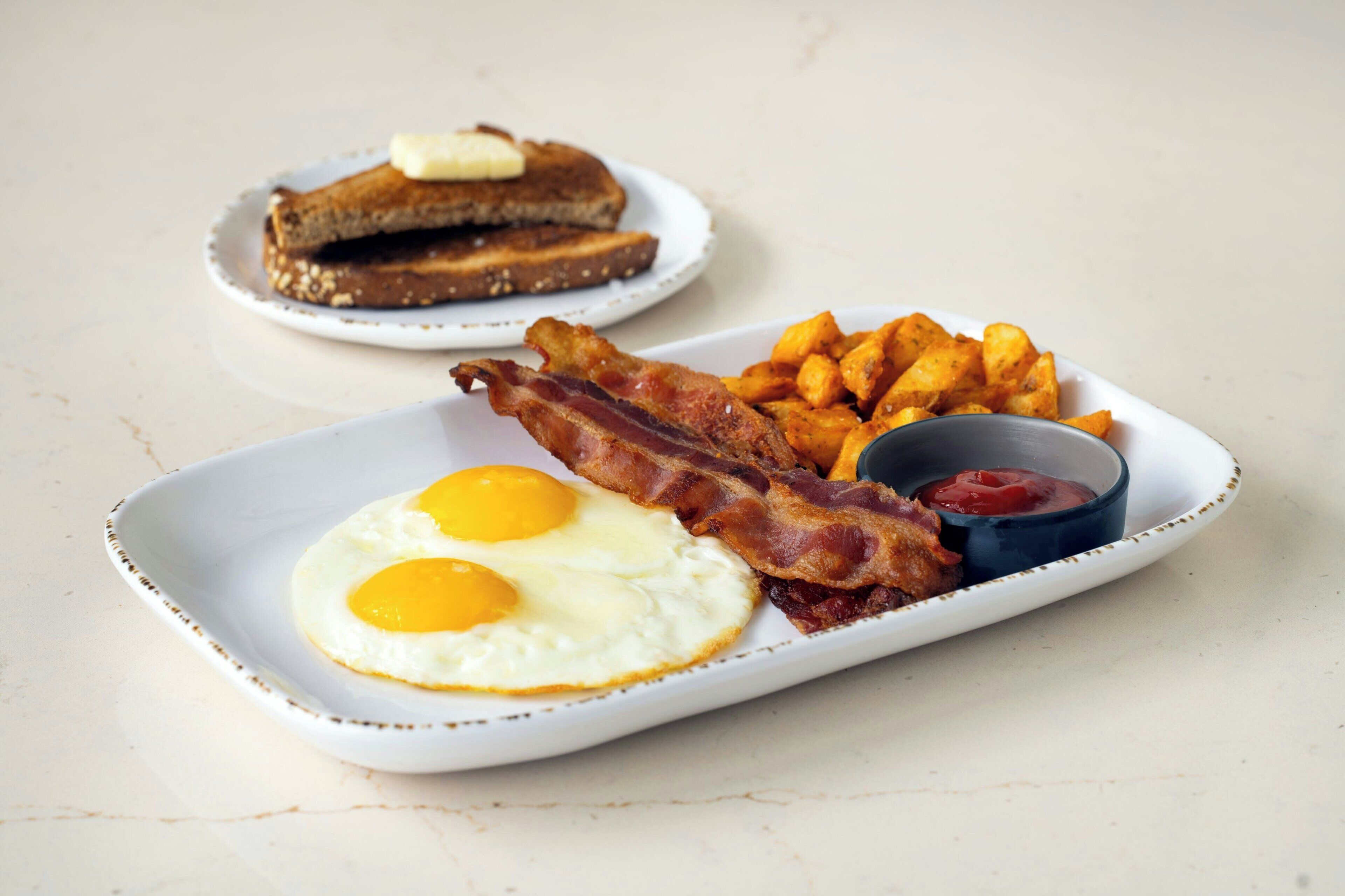 A breakfast plate with eggs, bacon, hash browns, toast, and ketchup.