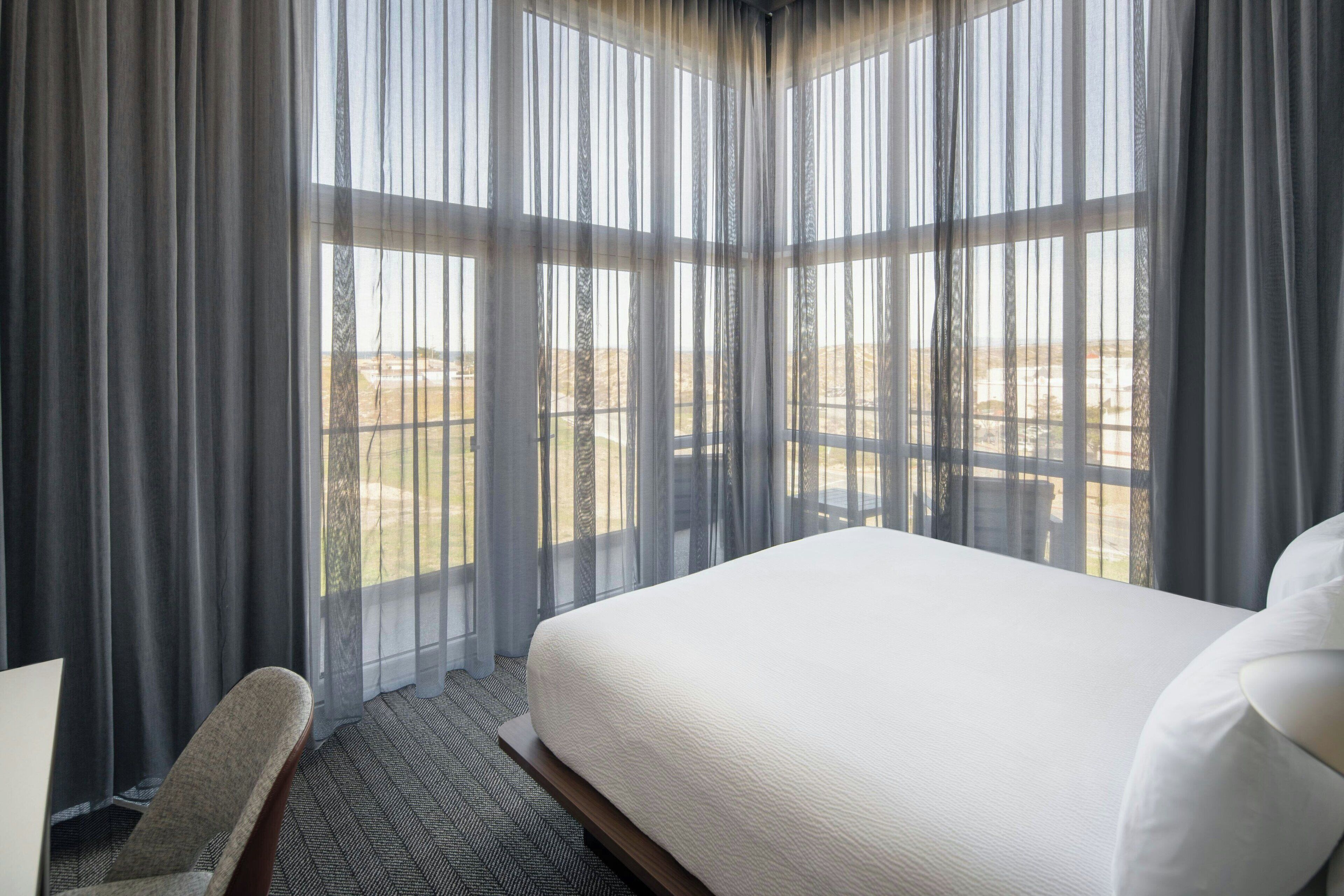 Habitación de hotel con una cama grande, una silla y vistas a un paisaje desértico a través de cortinas traslúcidas.
