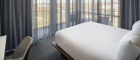 Habitación de hotel con una cama grande, una silla y vistas a un paisaje desértico a través de cortinas traslúcidas.