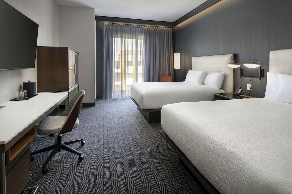 Une chambre d’hôtel équipée d’un bureau, de deux lits, d’une télévision et d’une grande fenêtre avec des rideaux.