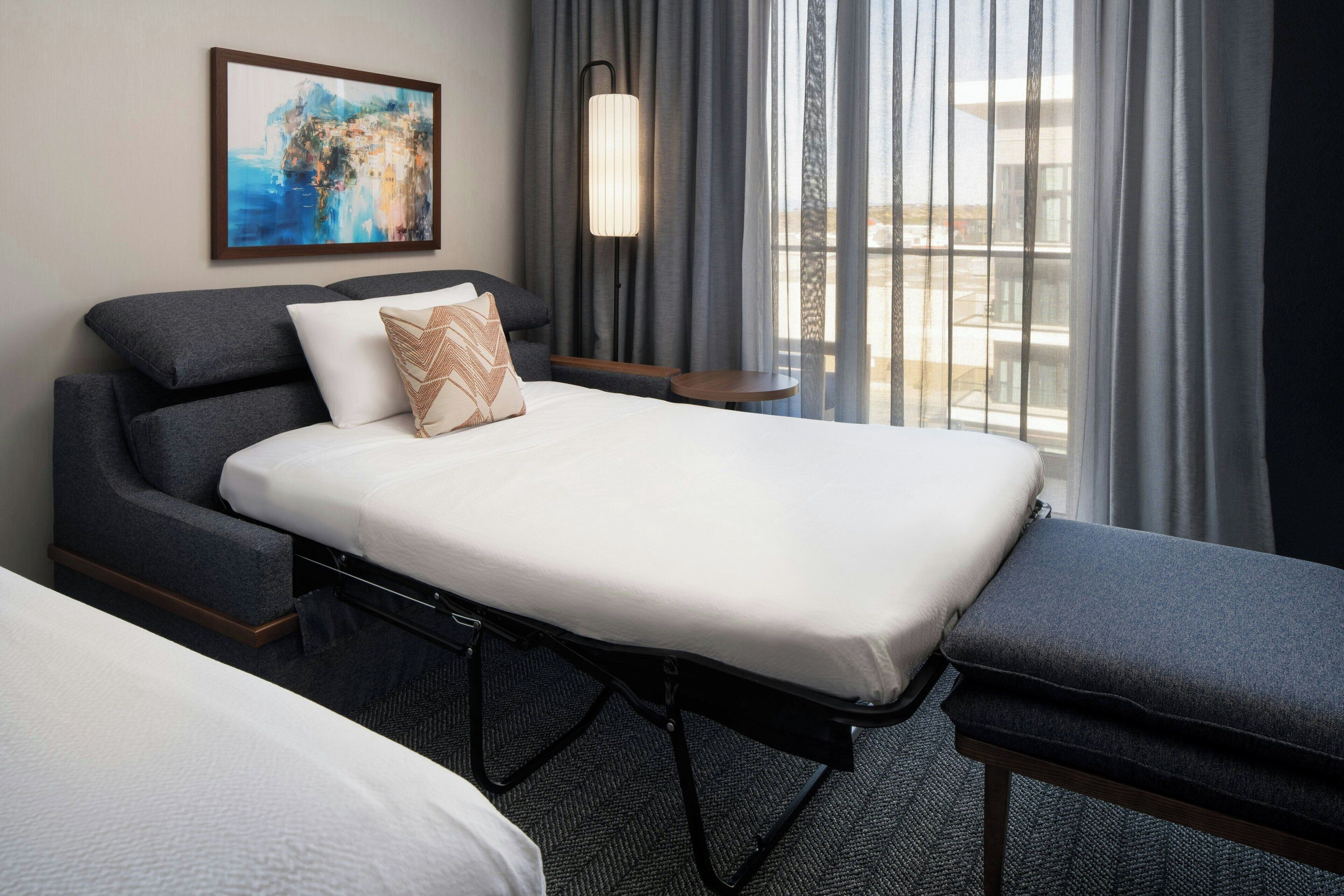 A hotel room with a bed, a bench, a bedside table, a lamp, and a painting on the wall.