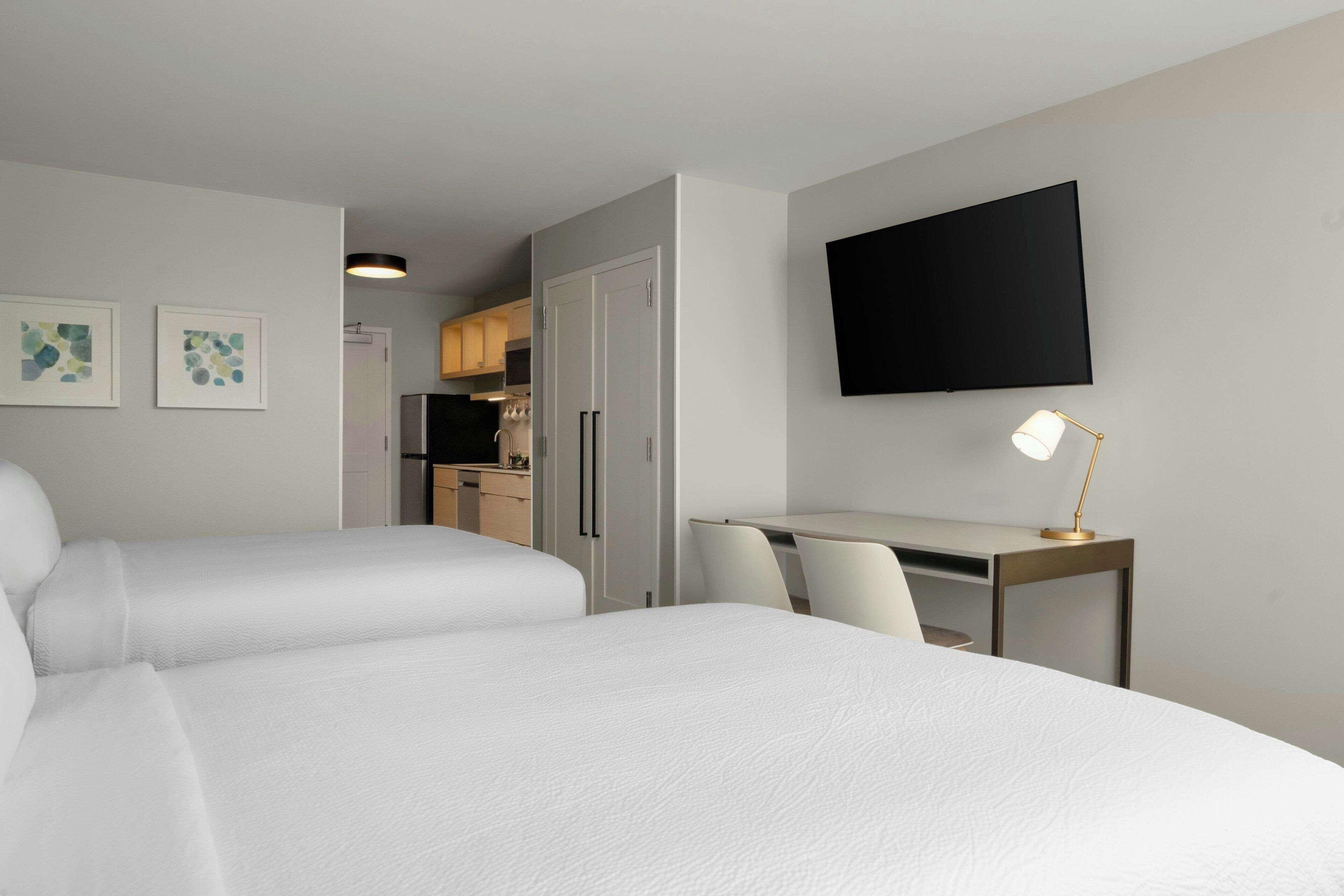 Ett hotellrum med två sängar, ett skrivbord, en stol, en tv och ett litet kök i bakgrunden.