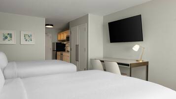 Ett hotellrum med två sängar, ett skrivbord, en stol, en tv och ett litet kök i bakgrunden.