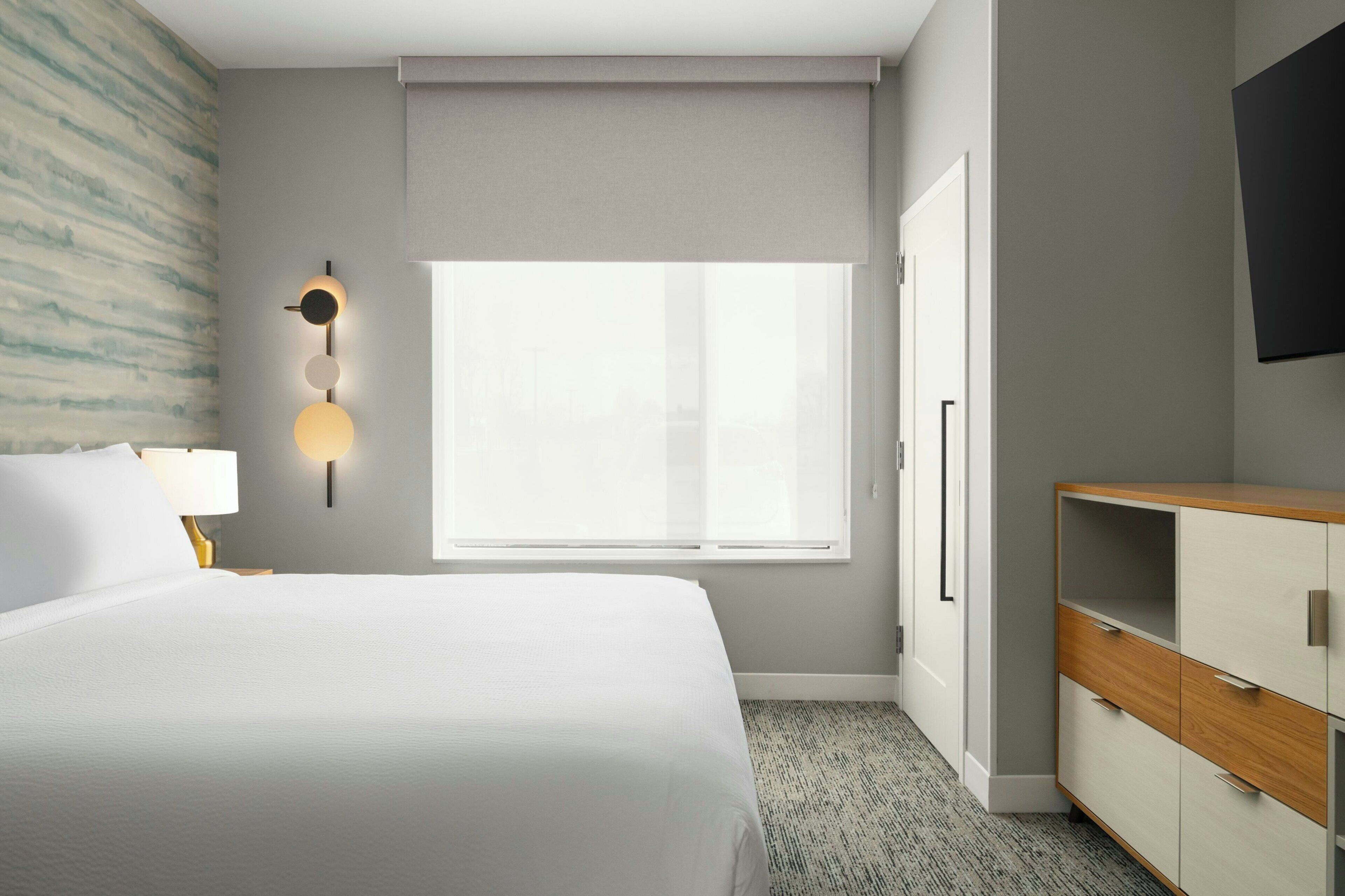 A hotel room with a bed, a desk, a chair, a window, and a TV mounted on the wall.