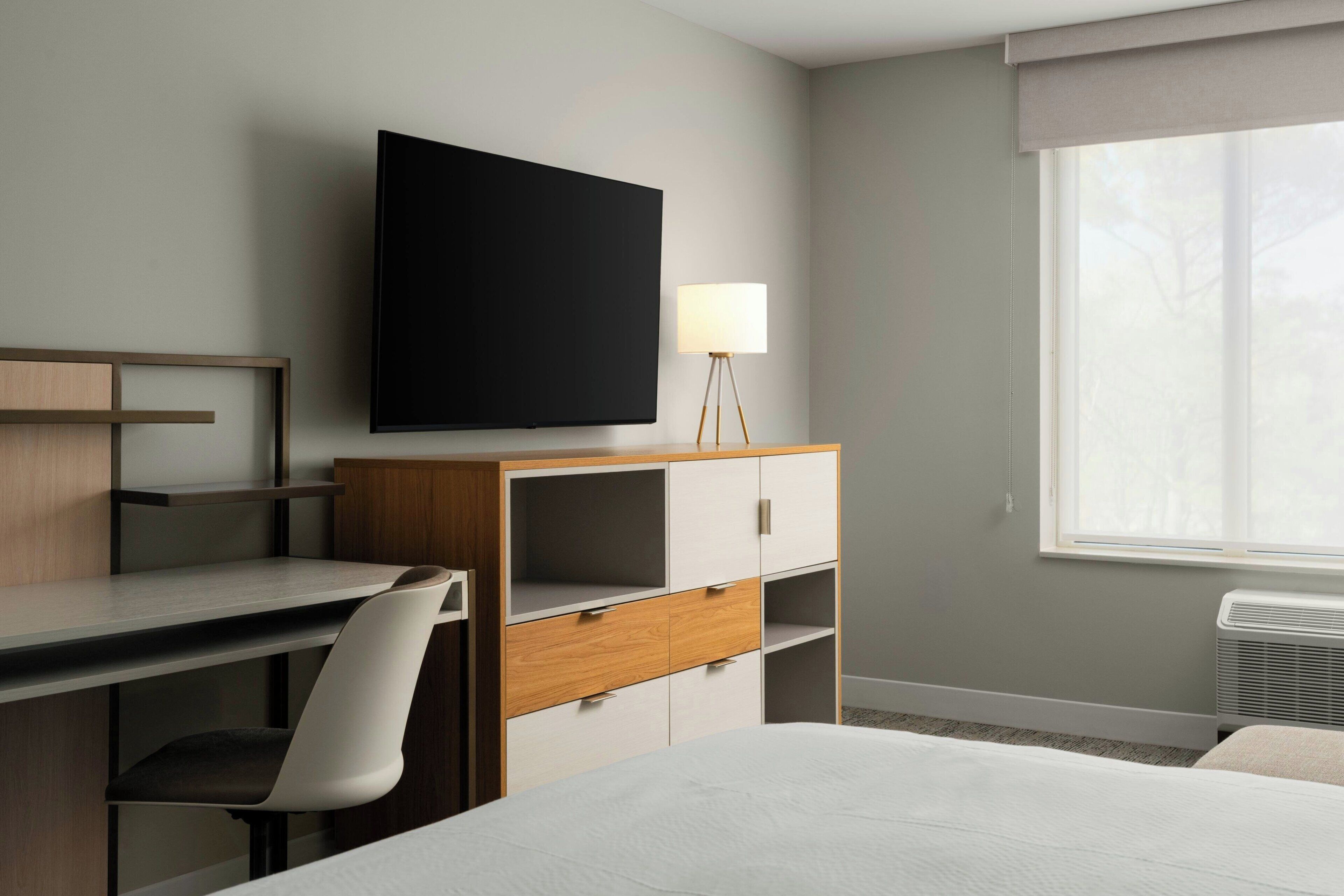 A hotel room with a bed, a desk, a chair, a television, a lamp, and a window.