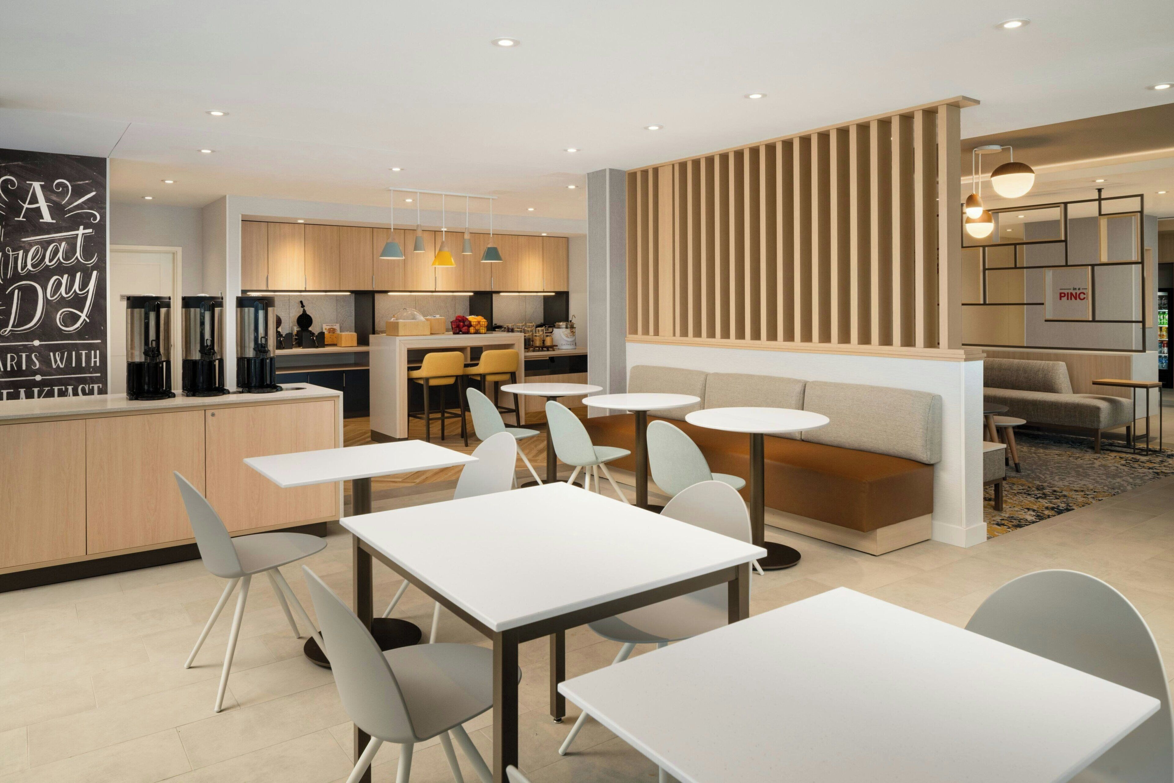 A modern cafe with white tables and chairs, a kitchen area, and a seating area with a wooden partition.