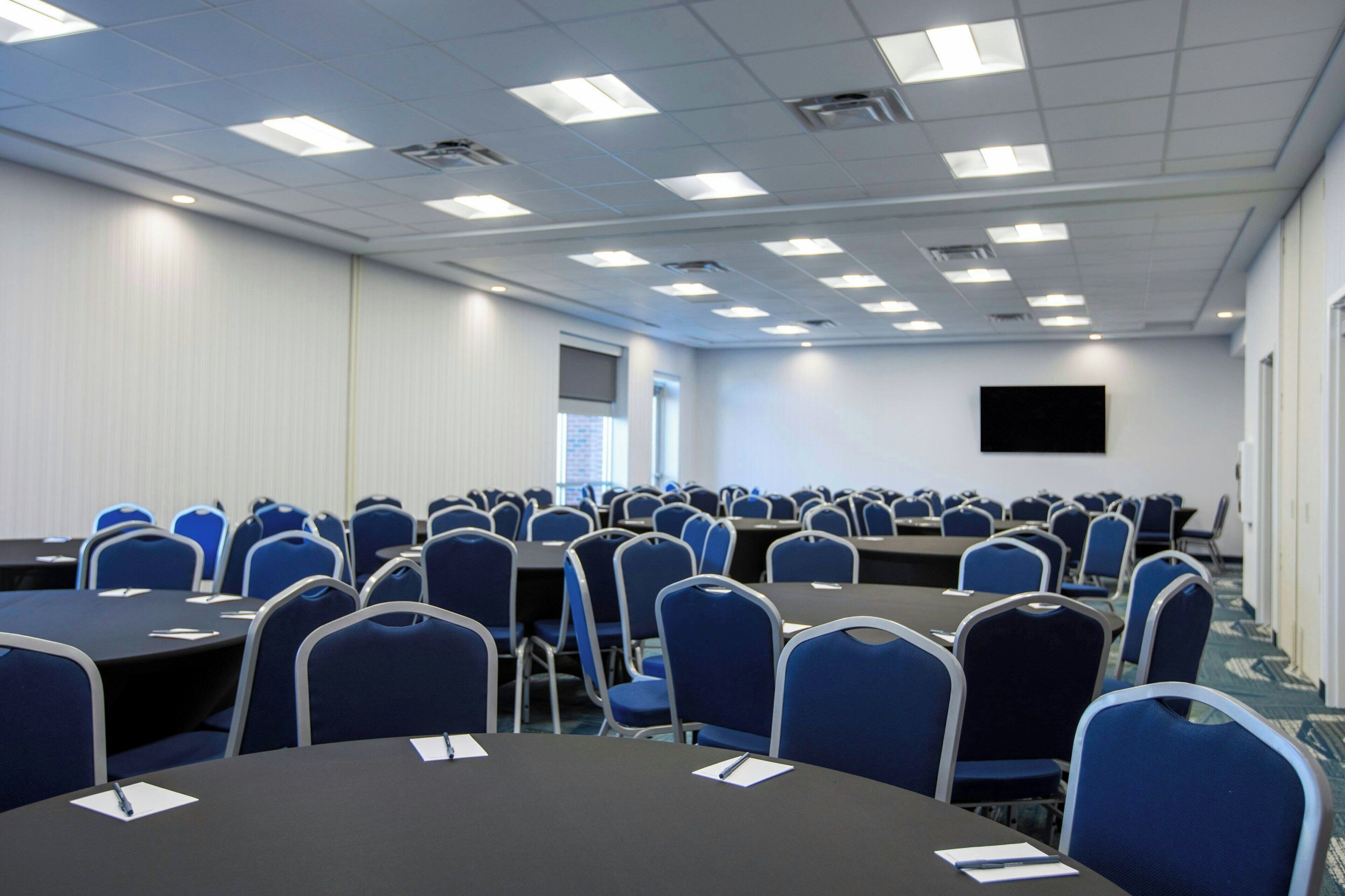 A conference room with multiple tables and chairs, a flat-screen TV on the wall, and a window with blinds.