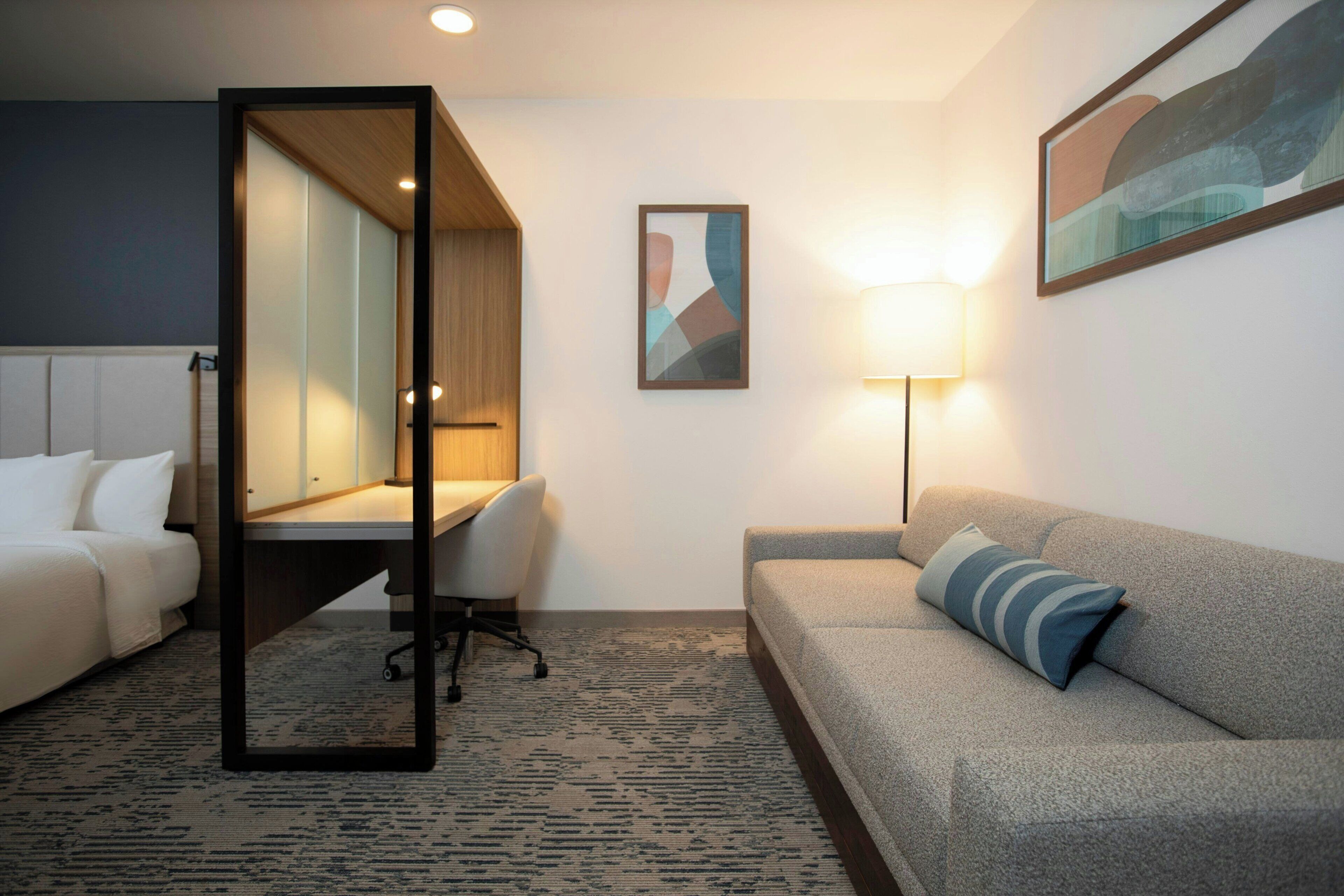 A hotel room with a bed, a desk with a chair, a sofa, a standing lamp, and a painting on the wall.