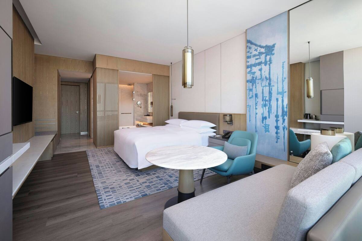 A hotel room with a bed, a desk, a chair, a sofa, a TV, and a large painting.