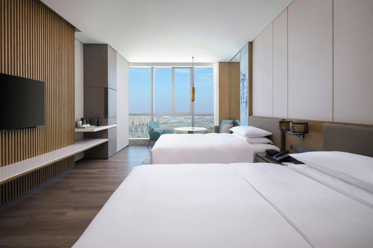 A hotel room with two beds, a TV, a desk, and a balcony with a city view.