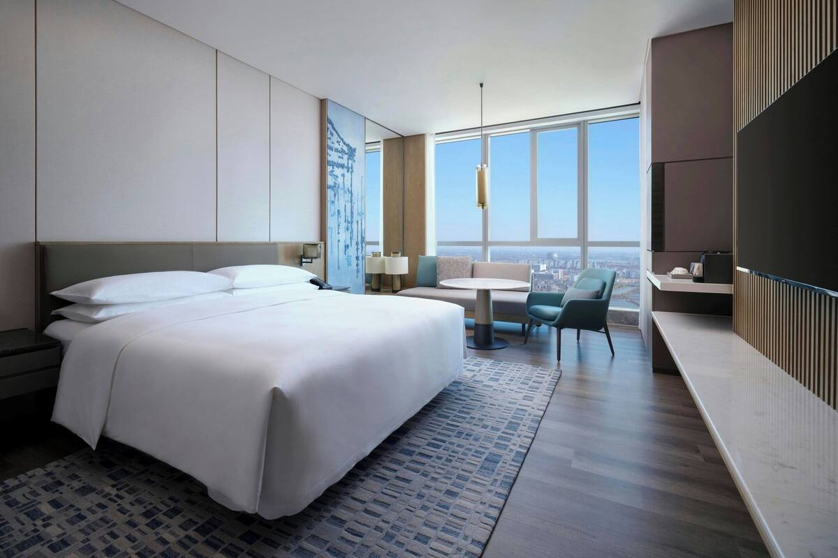A modern hotel room with a large bed, a desk, a chair, and a large window offering a city view.