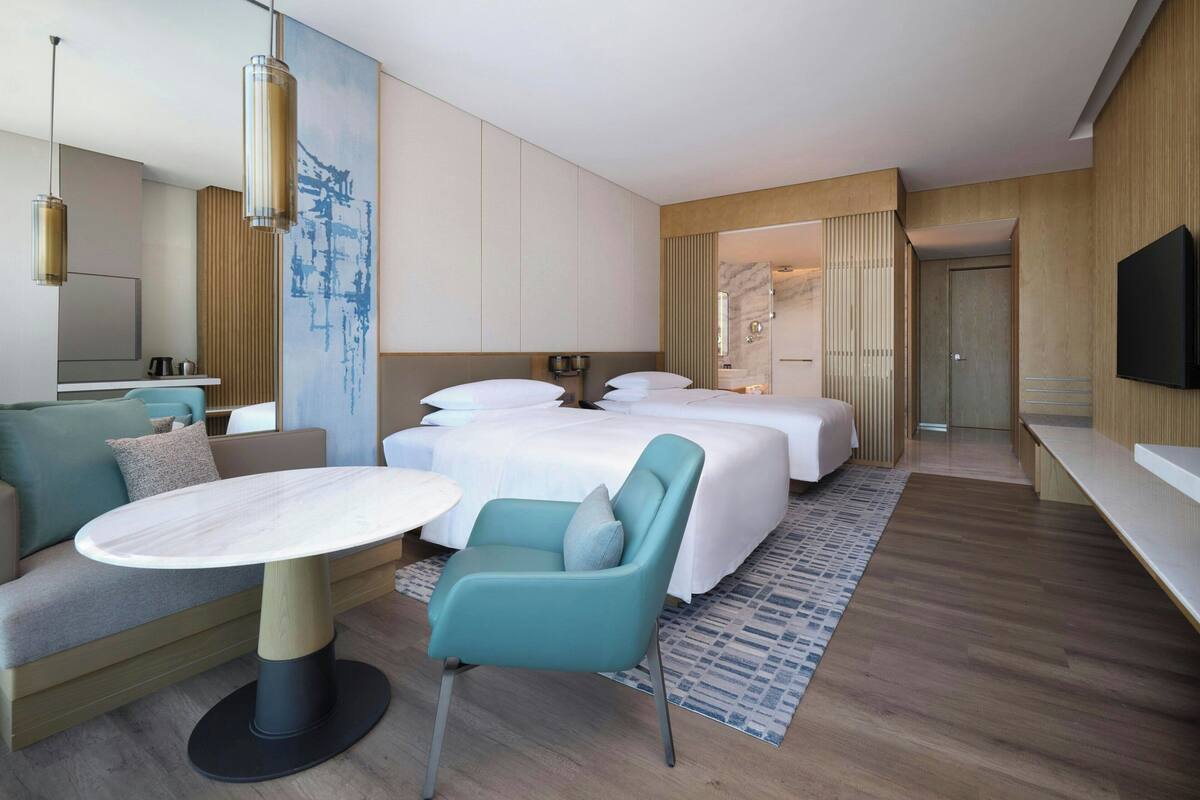 A modern hotel room with a sofa, two beds, a TV, and a large artwork on the wall.