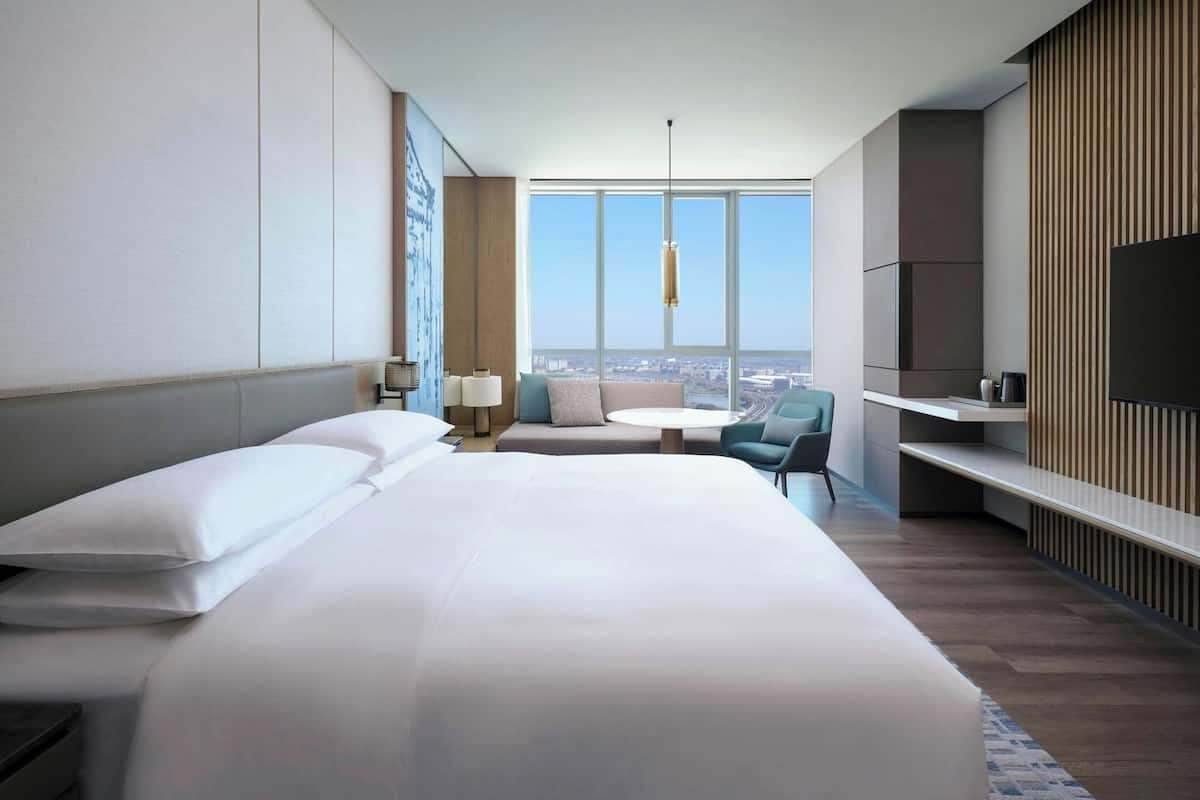 A hotel room with a large bed, a seating area with a round table, a TV on a stand, and a city view through large windows.