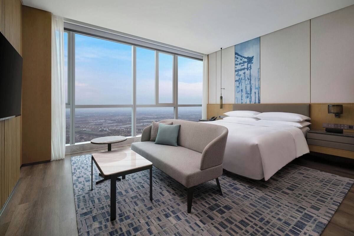 A hotel room with a large window, a sofa, a bed, a small table, and a patterned rug.