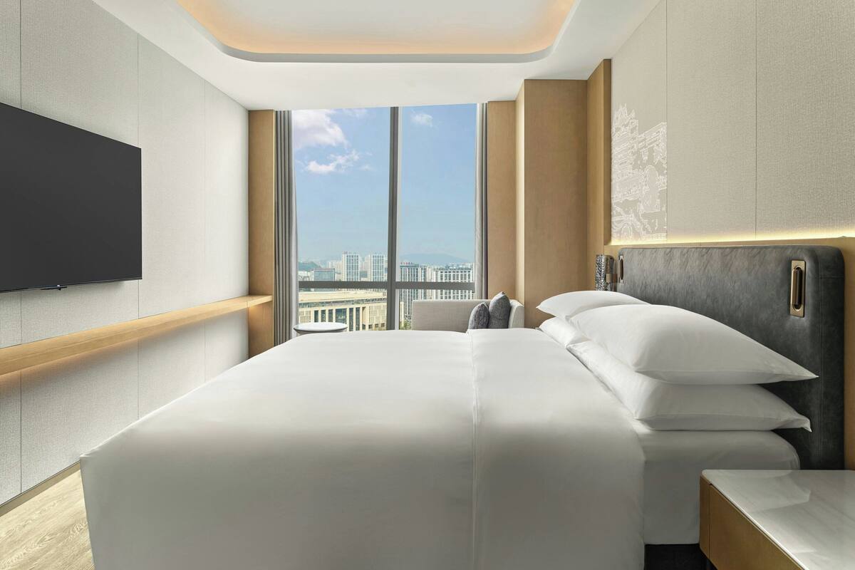 A modern hotel room with a large bed, a flat-screen TV mounted on the wall, and a city view through the window.