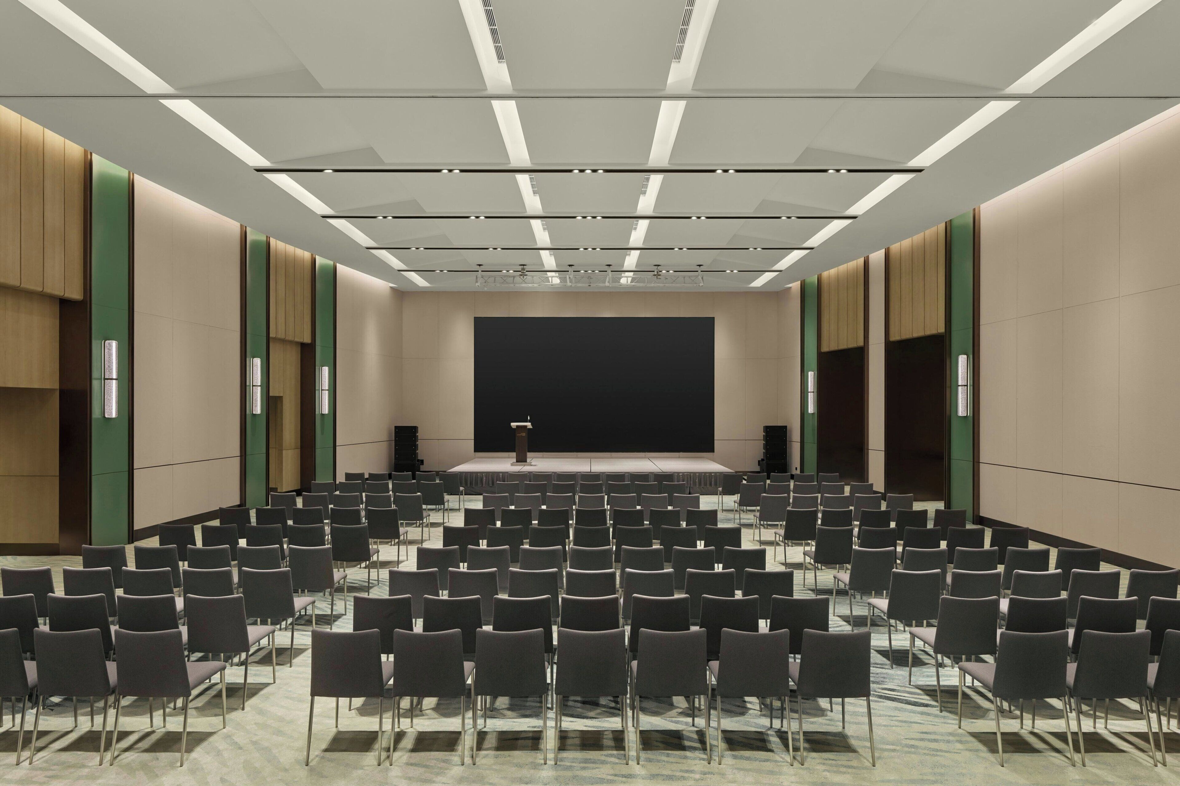 A conference room with rows of chairs, a stage with a podium, and a large screen.