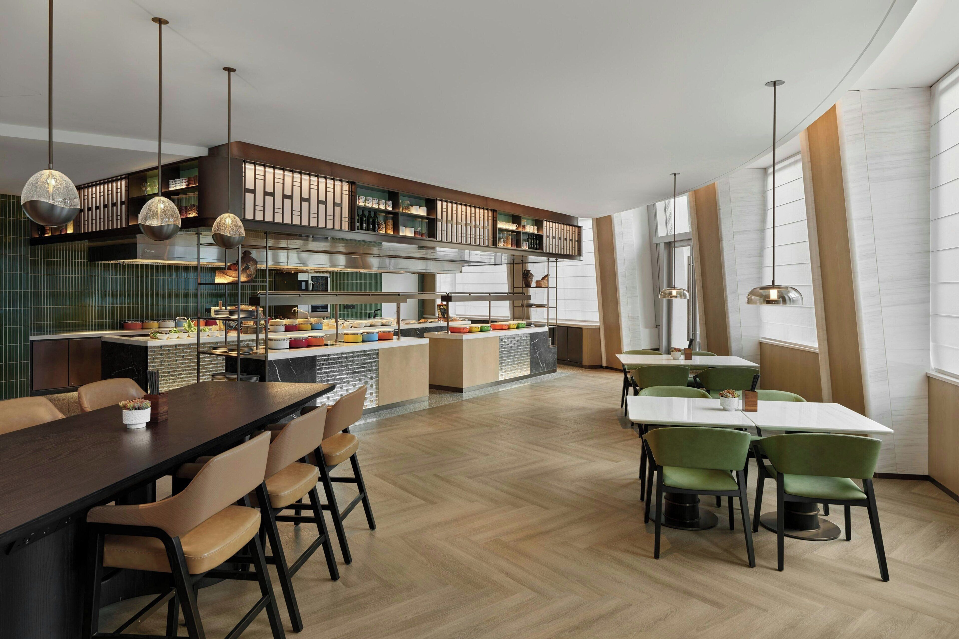 A modern restaurant with a long communal table, individual seating, and a kitchen area.