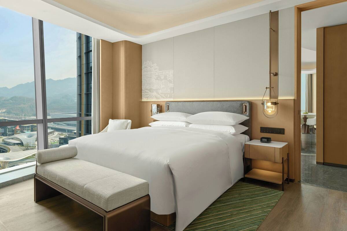A modern hotel room with a large bed, a bench, a nightstand, and a view of a cityscape.