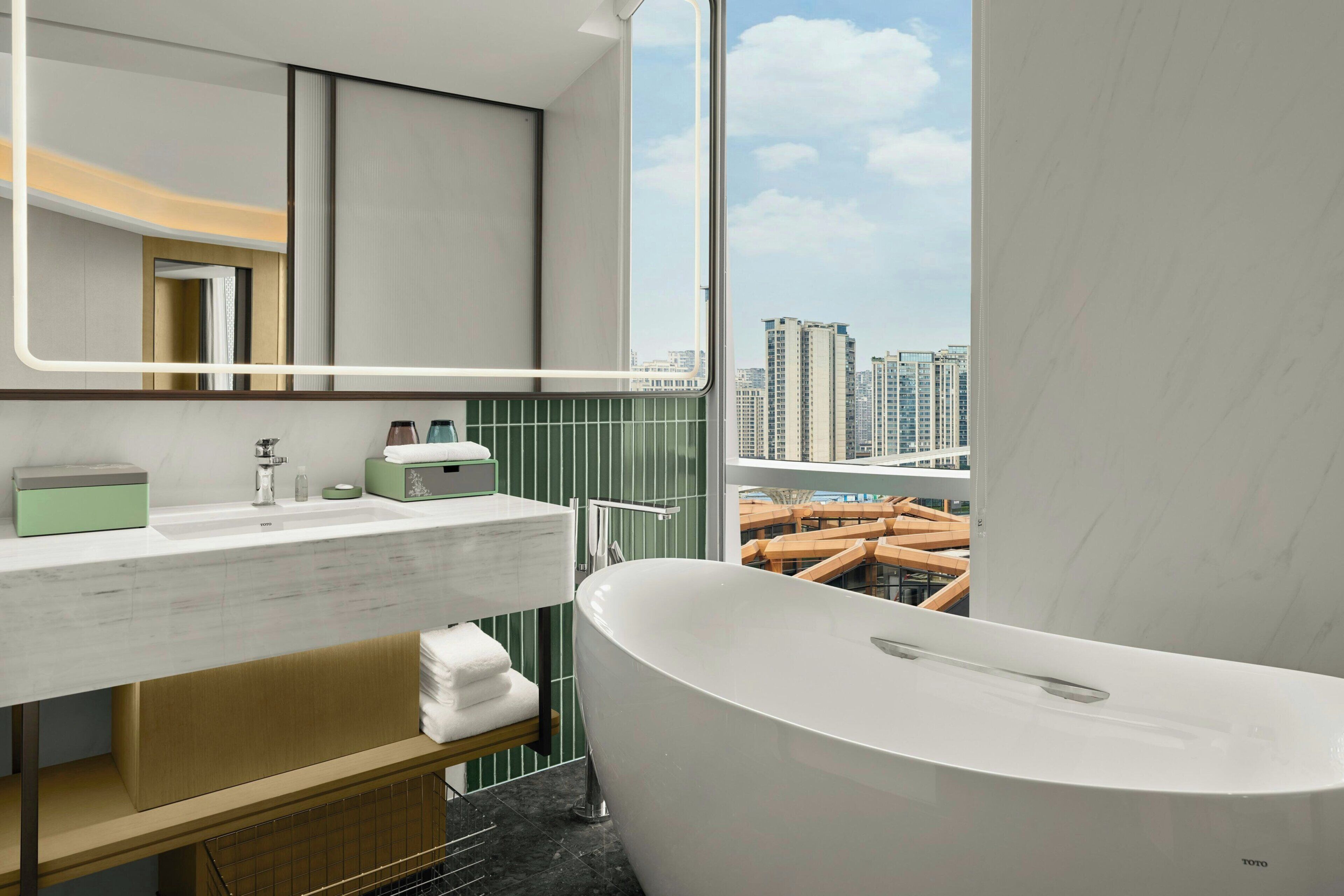 A modern bathroom with a large bathtub, a vanity with a mirror, and a view of a cityscape through a large window.