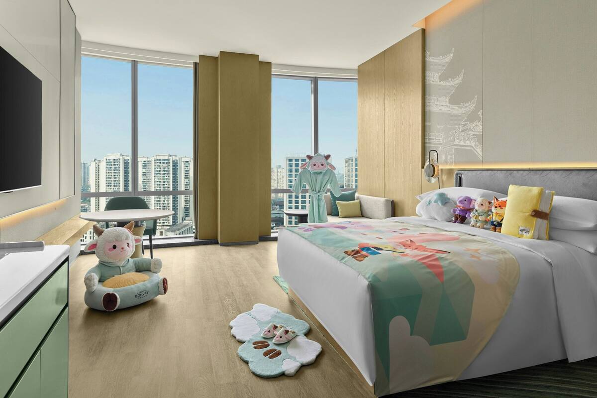 A modern hotel room with a large bed, a flat-screen TV, a dining area, and a view of high-rise buildings.