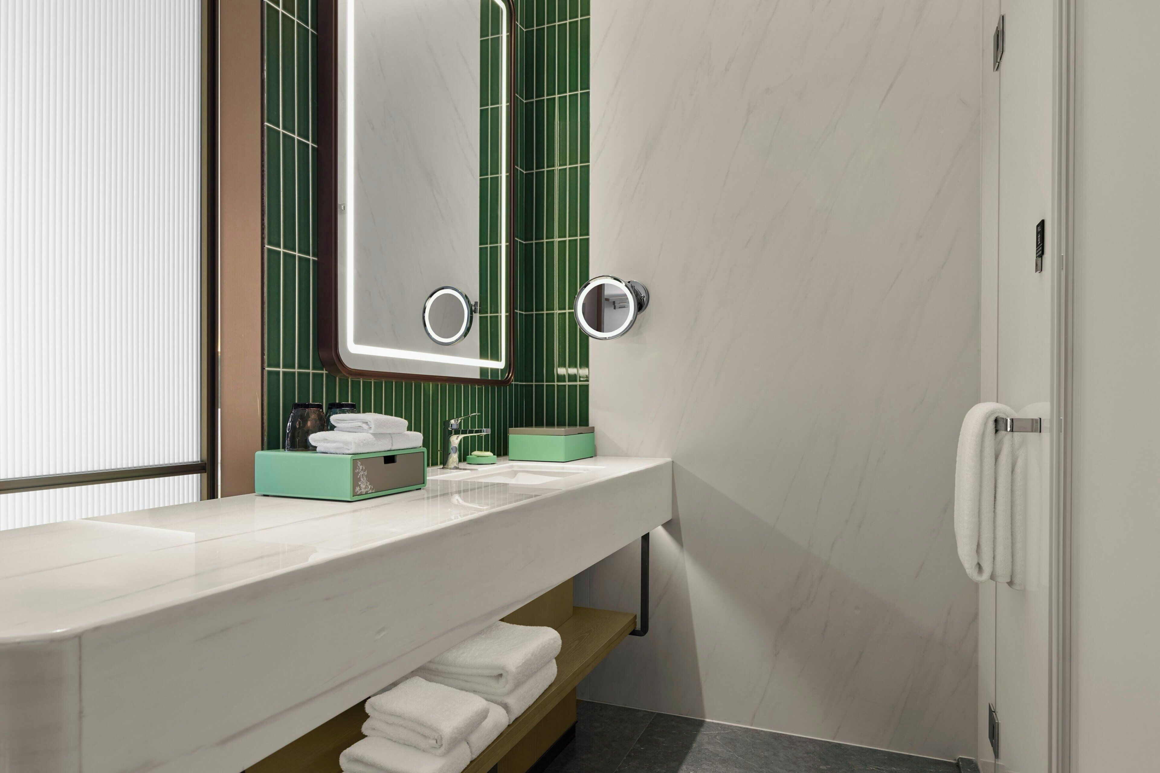 A modern bathroom with a white sink, green-tiled wall, and a mirror with built-in lights.