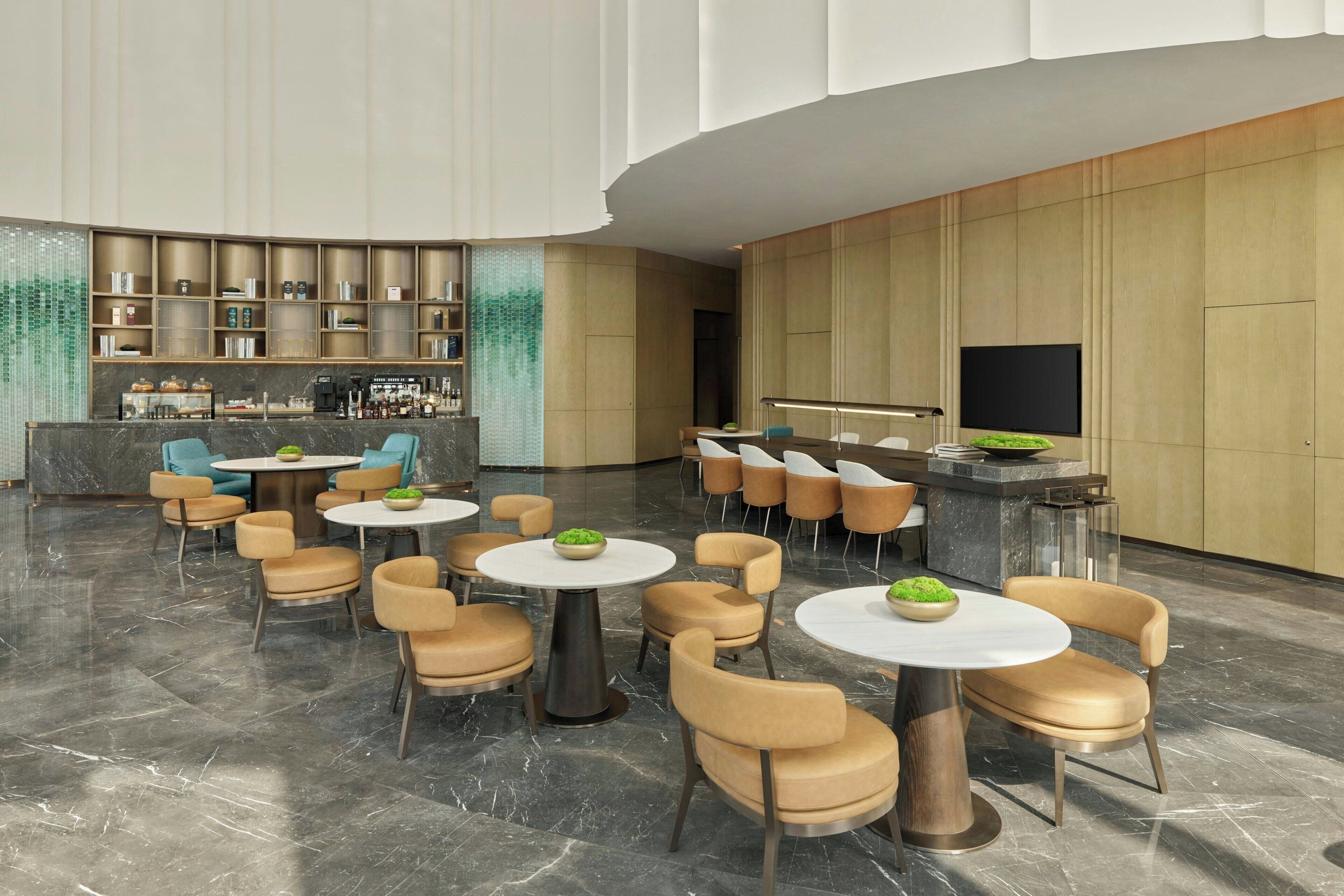 A modern lobby with round tables, comfortable seating, a bar area, and a television screen.