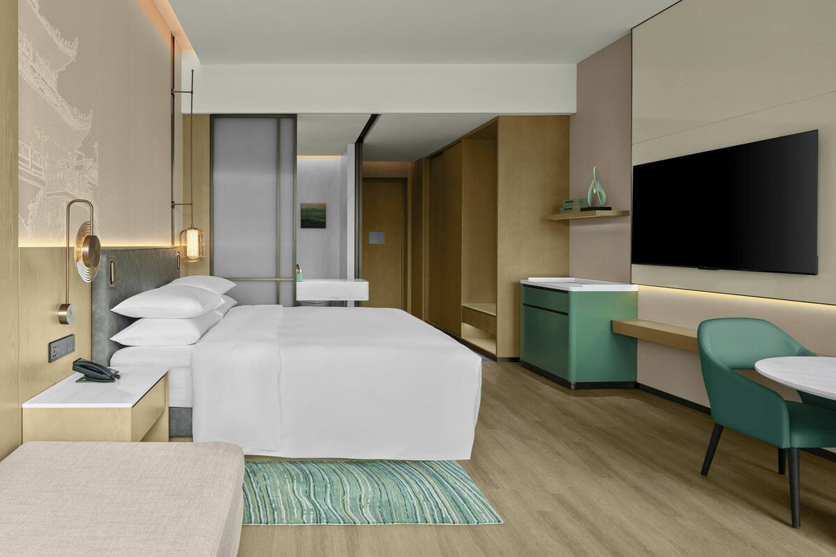 A modern hotel room with a bed, bedside table, lamp, TV, and a small dining area with a chair and table.
