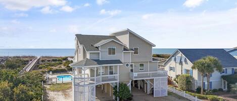 A two-story house with a covered porch and a pool, located near a beach with a clear sky.