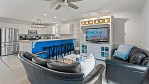 Una cocina moderna con una isla central, un comedor con taburetes de bar, una sala de estar con un sofá de cuero negro y un televisor en la pared mostrando 'ROACTIVE VACATIONS'.