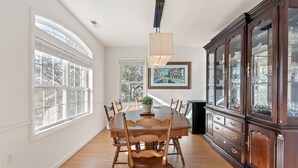 Un comedor con mesa y sillas de madera, un aparador grande con puertas de cristal y ventanas amplias que dan a vistas de árboles y edificios.