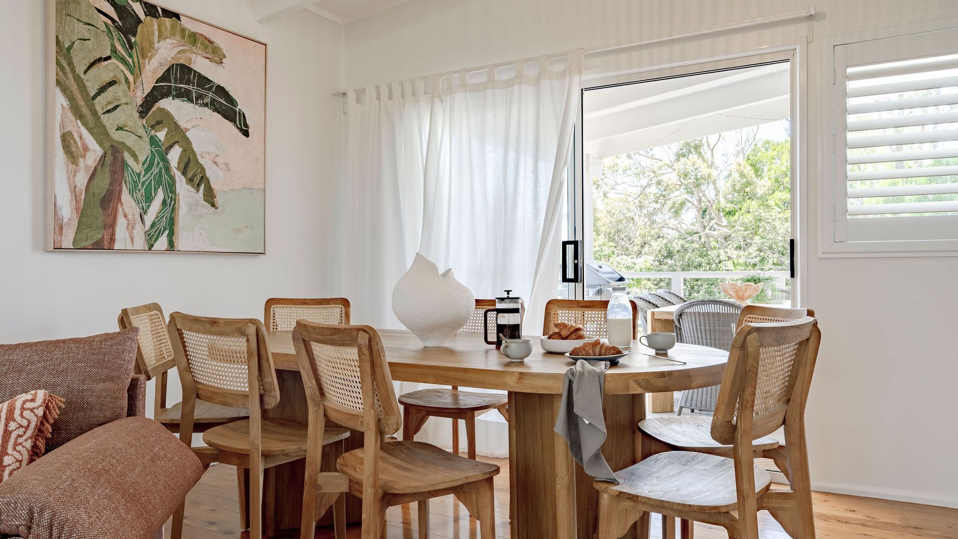 A dining area with a wooden table and chairs, a painting on the wall, and a view of greenery outside.