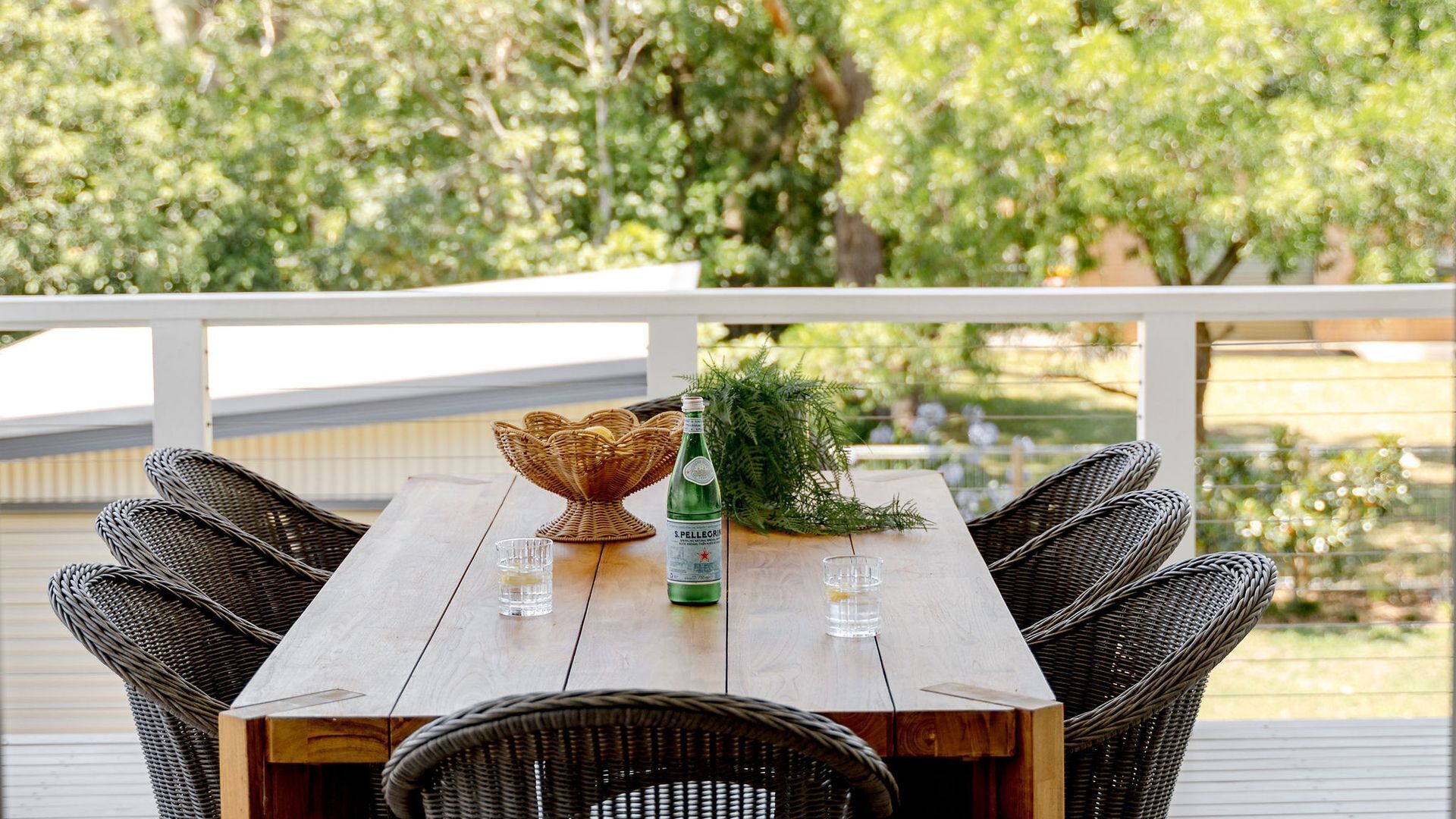 Une terrasse en bois avec des chaises en osier et une table, agrémentée d’une bouteille d’eau Pellegrino et d’un bol décoratif.