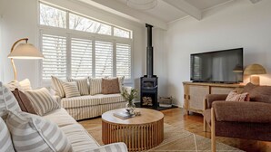 Ein gemütliches Wohnzimmer mit einer Couch, einem Sessel und einem Fernseher. Ein Kamin und ein großes Fenster mit Jalousien sind ebenfalls vorhanden.