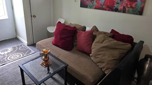 Un salón con un sofá color beige, cojines rojo y marrón, una mesa de centro de cristal con una vela, y un cuadro de flores rojas en la pared.