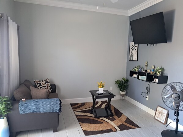 A living room with a grey sofa, a small table with a vase, a wall-mounted TV, and a fan.