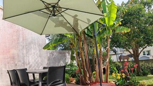 A patio with a table and chairs, a large umbrella, and a garden with various plants and flowers. - Residential neighborhood, pleasant south-facing house (FOURAS)