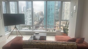 Een moderne woonkamer met een groot raam, een flatscreen televisie op een driepoot, een bank, een salontafel en een potplant.