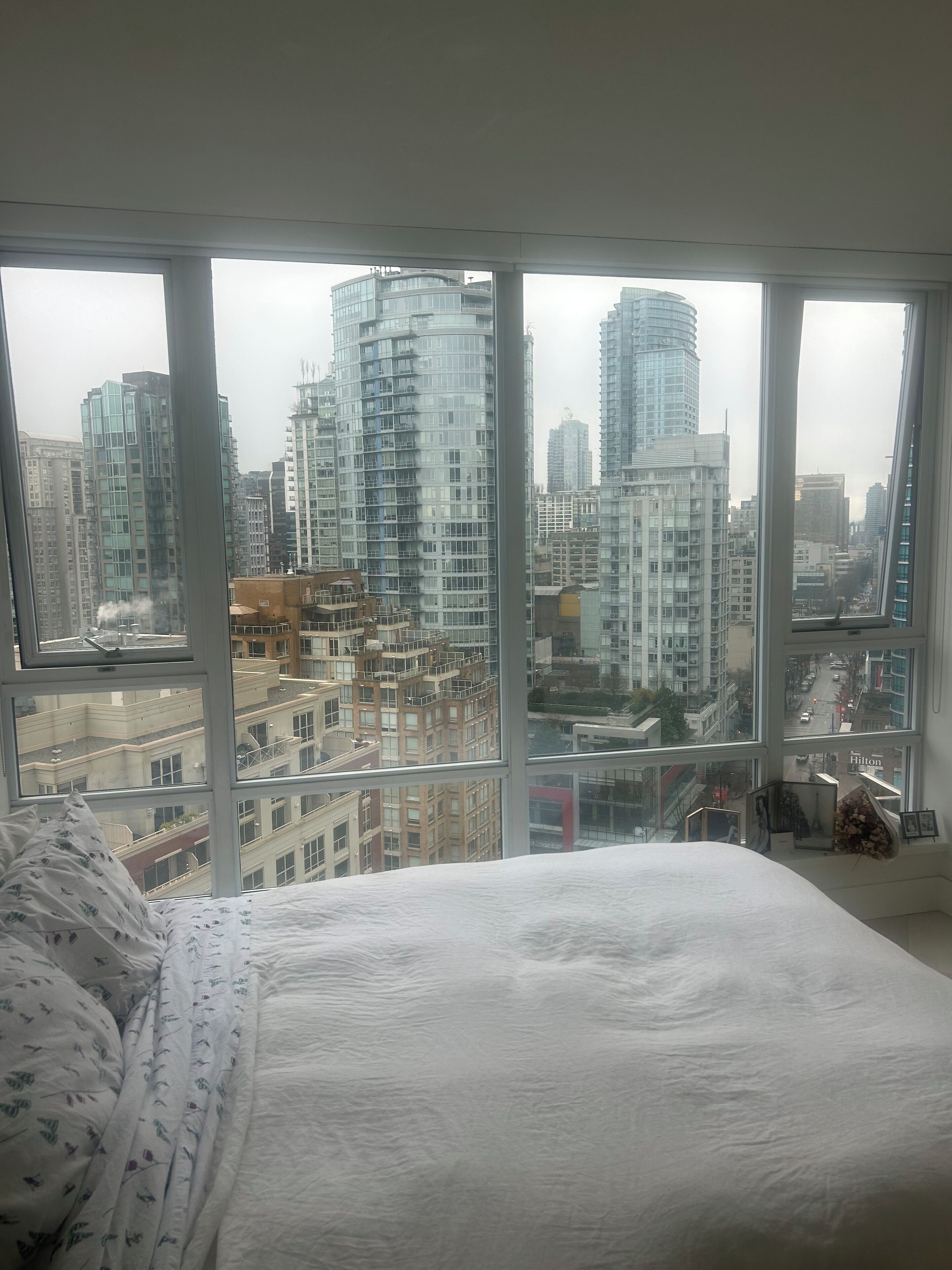 A bedroom with a large window offering a city view, a bed with patterned bedding, and a small table with personal items.