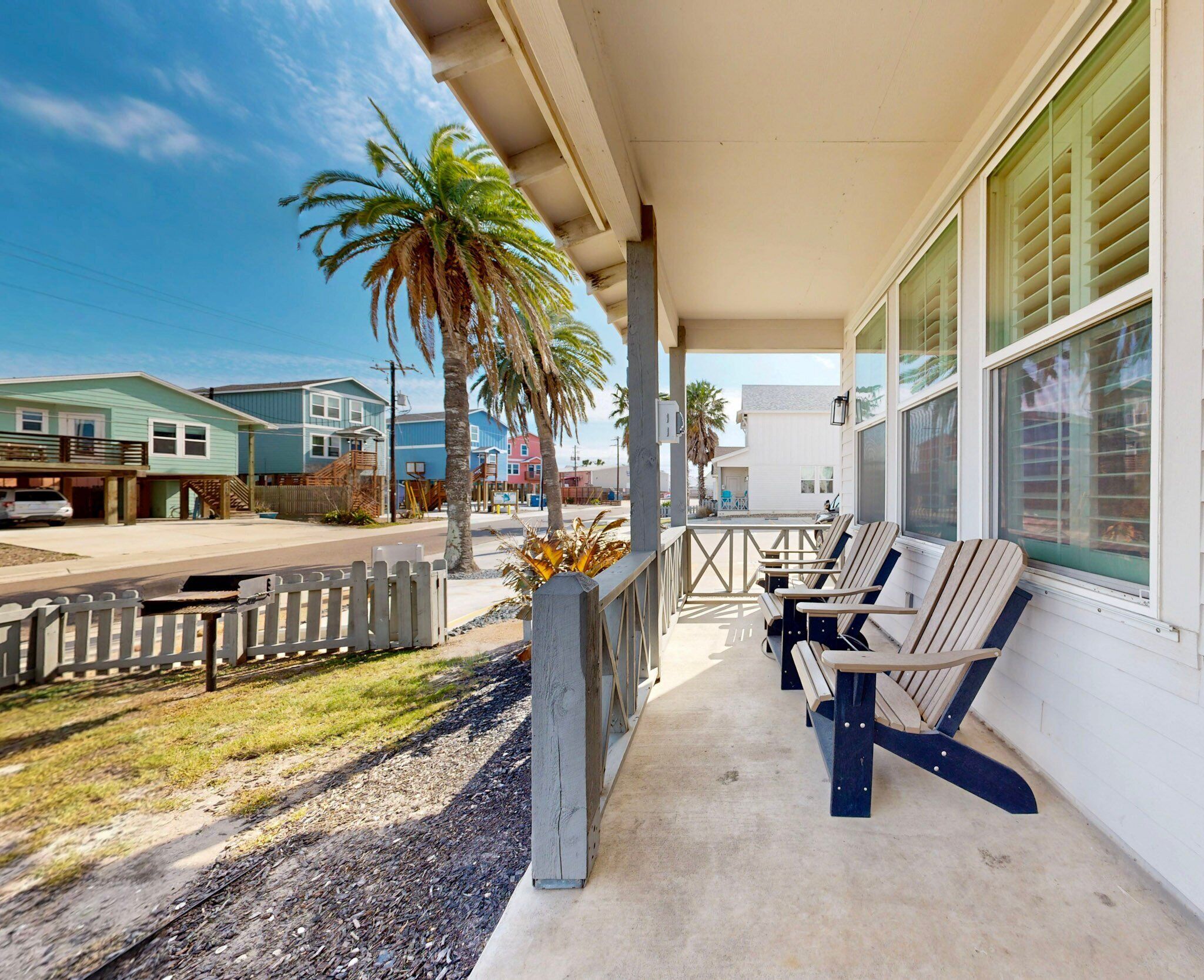 A covered porch with outdoor seating, a view of a street with houses, and a palm tree.