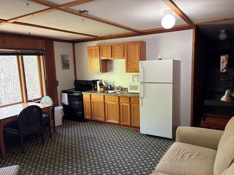 A compact kitchen with wooden cabinets, a white refrigerator, a stove, and a microwave. There is a dining area with a table and chairs, and a patterned carpet.. Spacious, comfortable 1 BR with FULL kitchen, large sitting rm, private bath