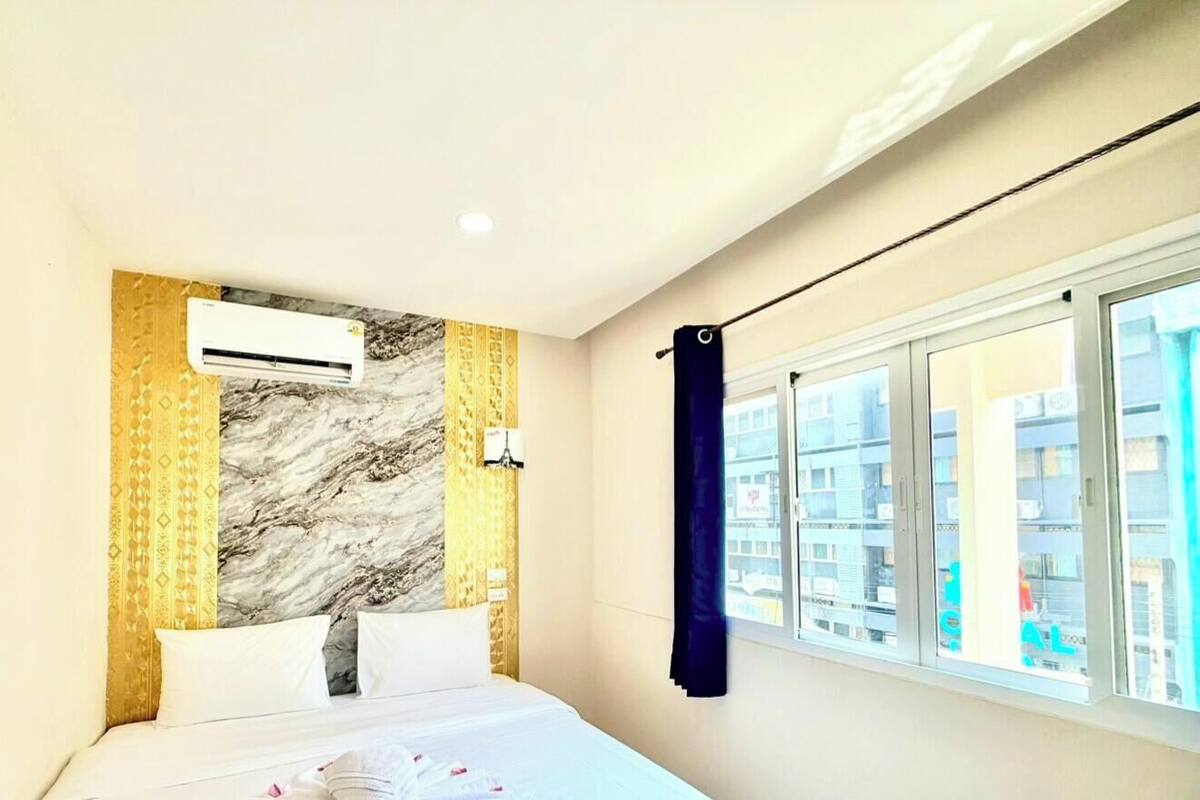 A hotel room with a bed, an air conditioning unit, a window with a curtain, and a view of buildings outside.