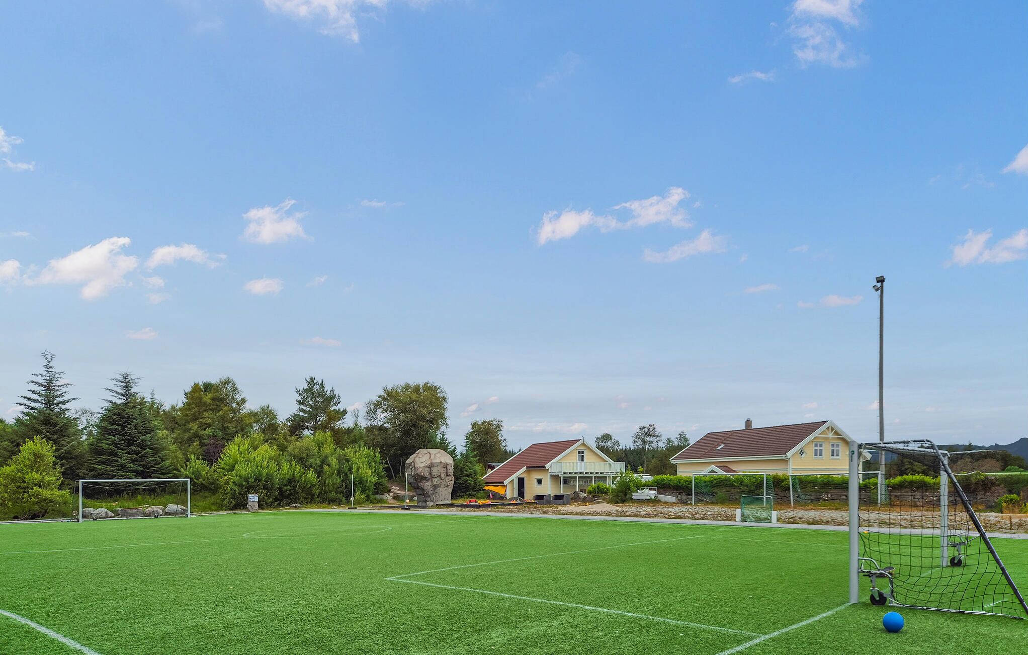 Um campo de futebol com um gol, uma escultura de pedra e casas ao fundo.