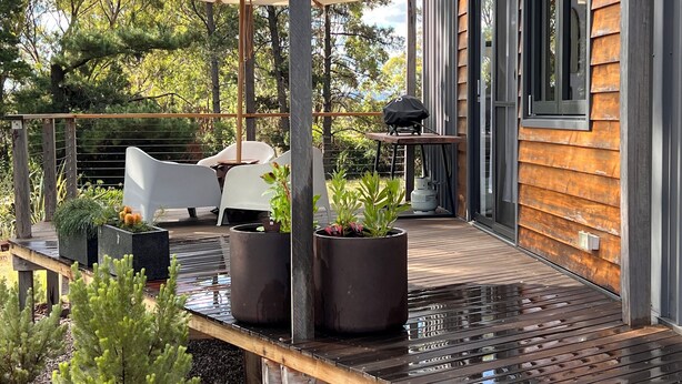 Een terras buiten met een houten terras, een tafel met stoelen, een barbecue en potplanten.