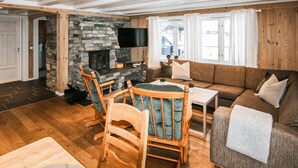 Ein gemütliches Wohnzimmer mit Kamin aus Stein, Holzmöbeln und einem großen Fenster mit Vorhängen.