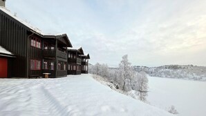 En flervåningsbyggnad i trä med balkonger, som ligger inbäddad i ett snötäckt landskap med utsikt över en sjö och träd.